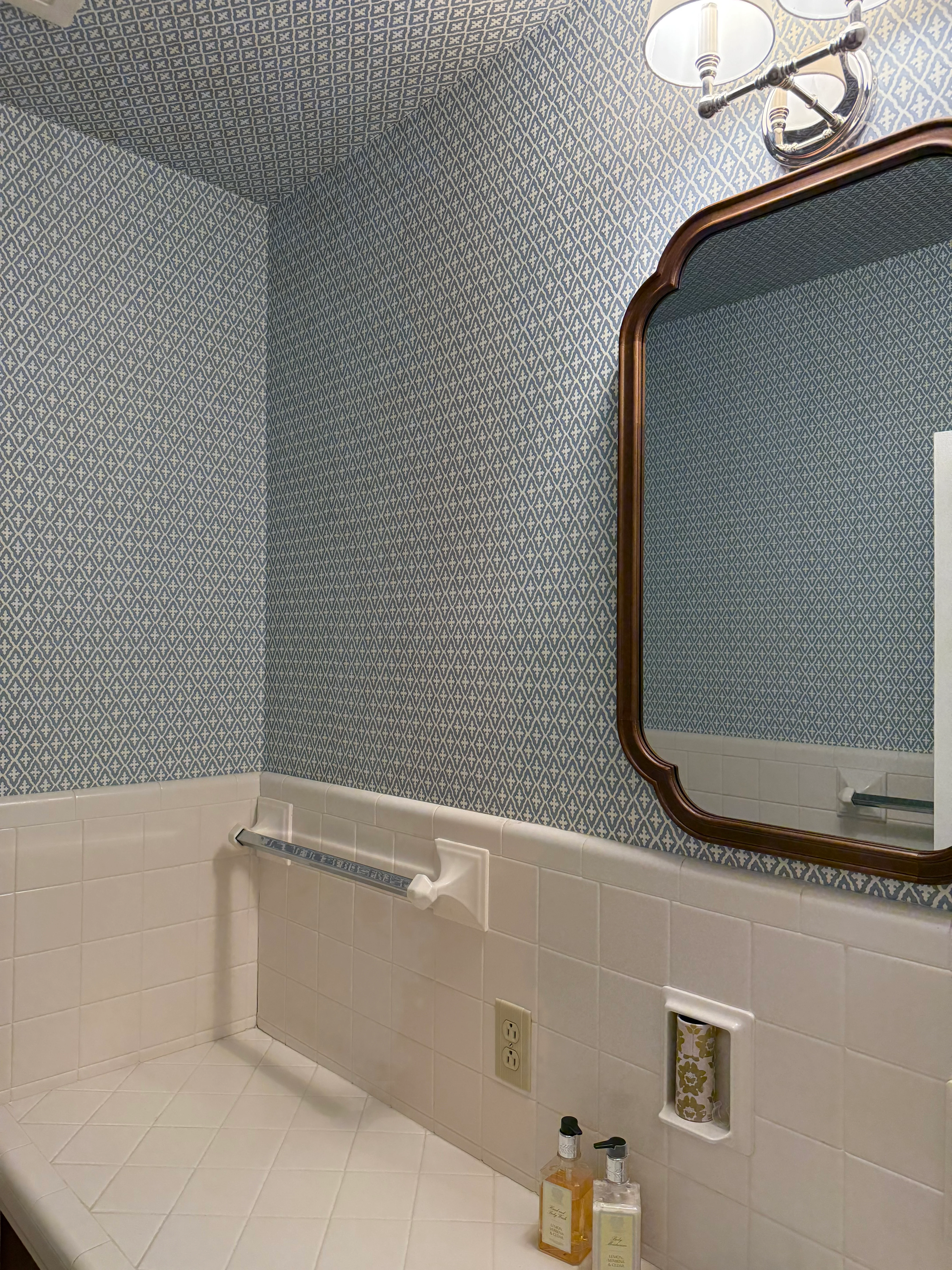 A powder room with a small blue diamond and fleur-de-lis pattern wallpaper above white tile wainscoting, with a wood-framed mirror.
