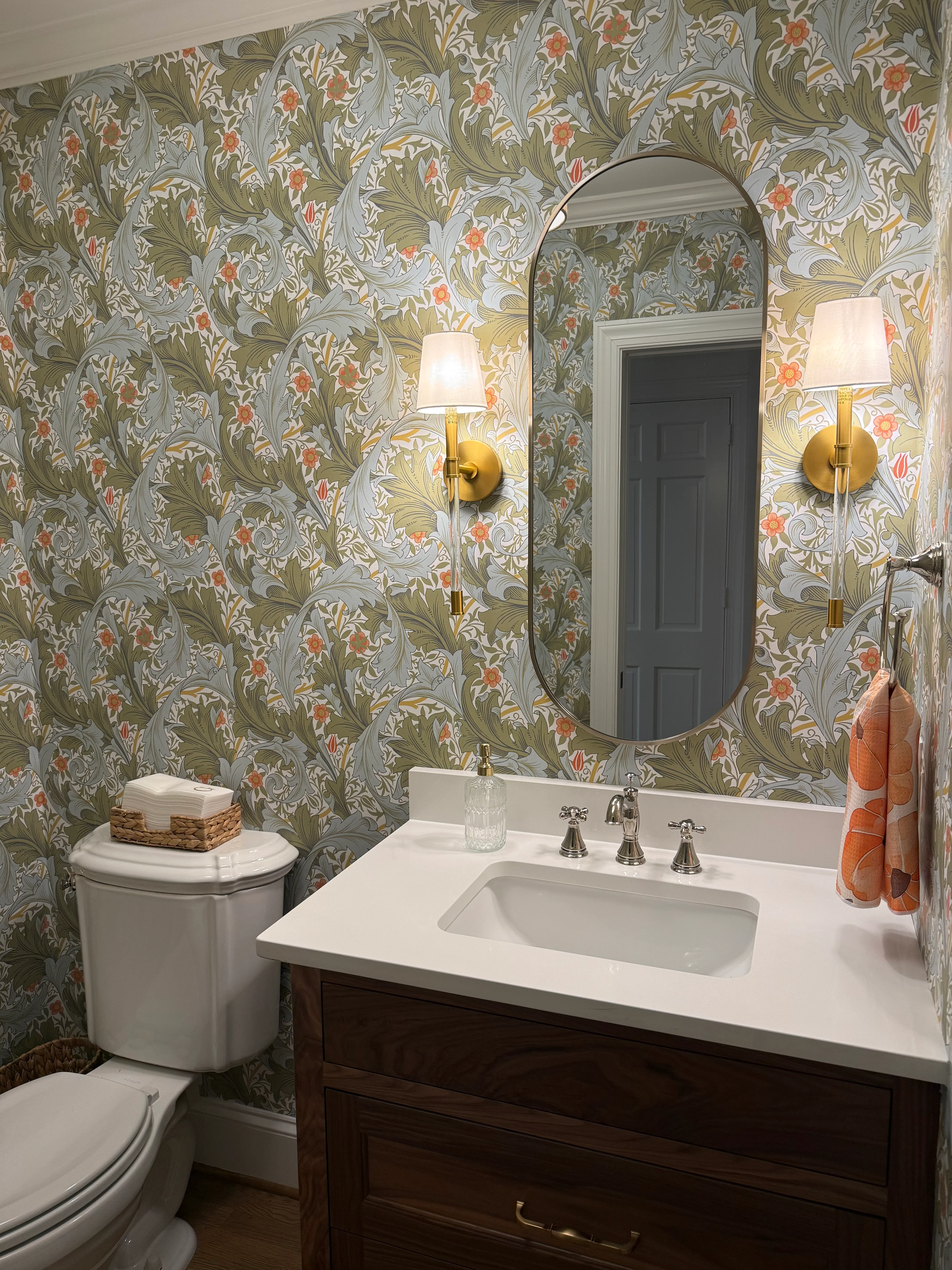 A powder room covered floor to ceiling in a dense acanthus leaf wallpaper in sage, slate, and coral, with brass sconces and an oval mirror.
