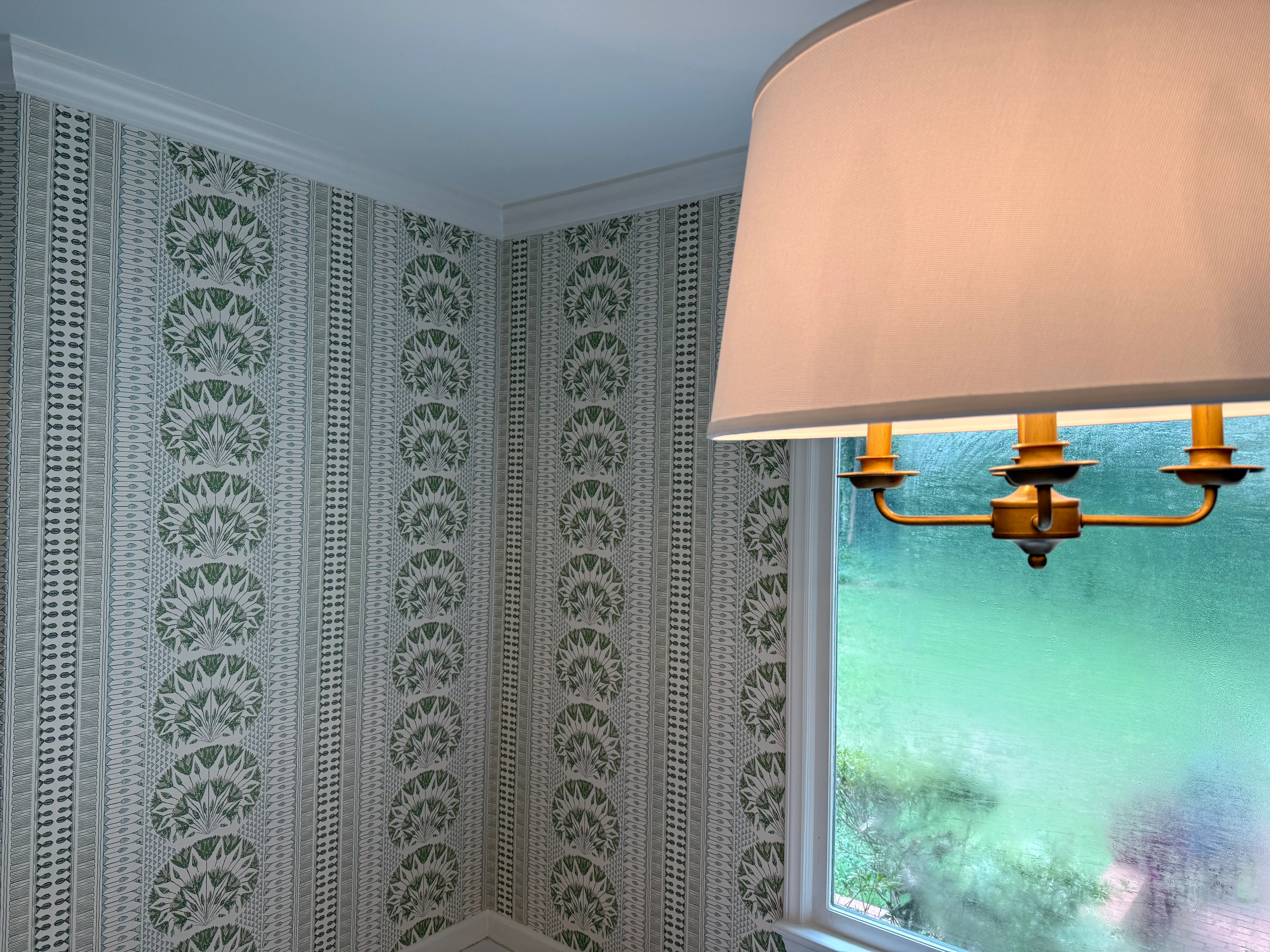 A room with a bold green vertical stalk and leaf pattern wallpaper against white trim, with a brass pendant shade visible.