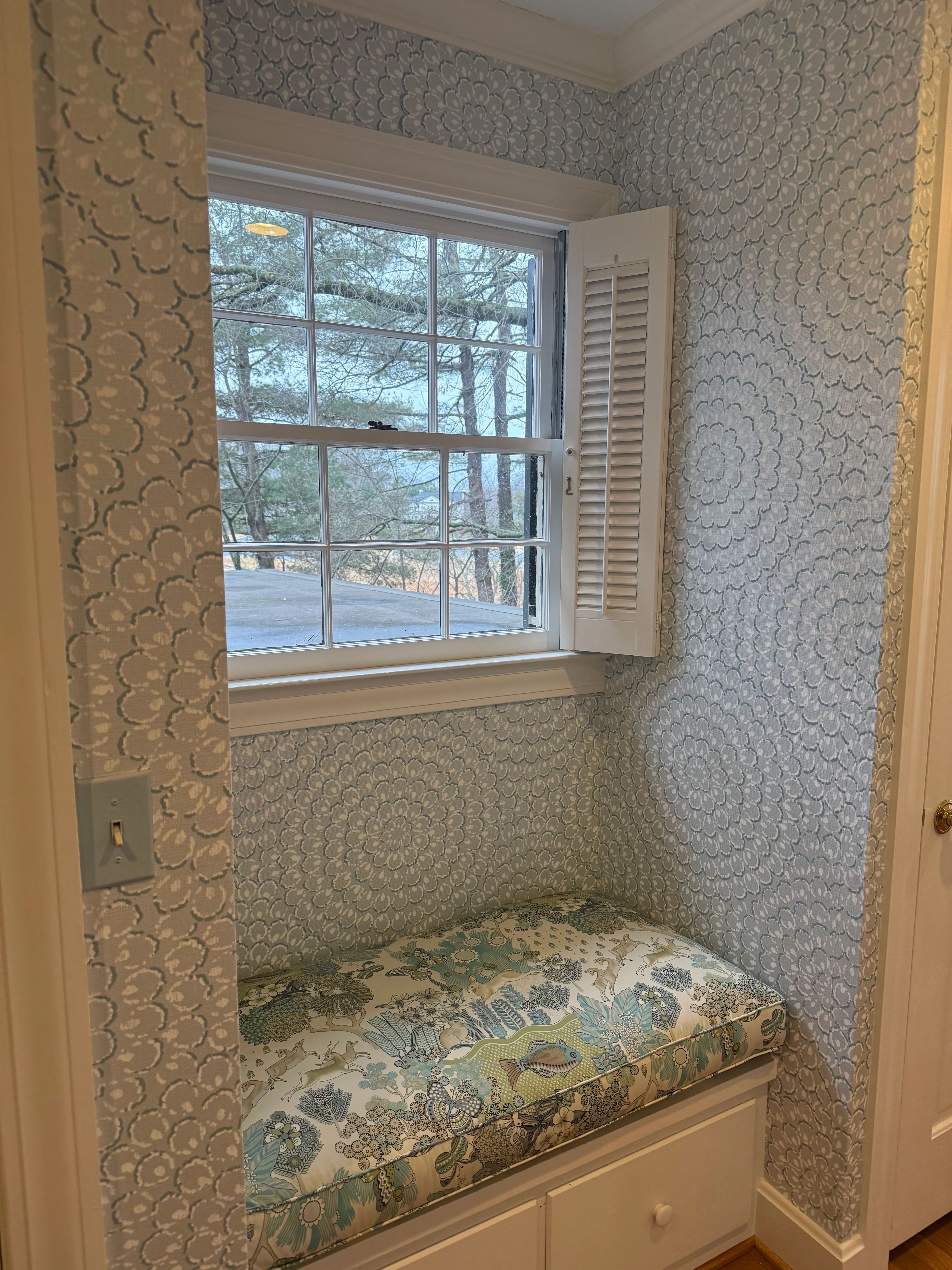 A window-seat nook wrapped in a soft grey-blue floral medallion wallpaper, with an upholstered cushion and natural light.