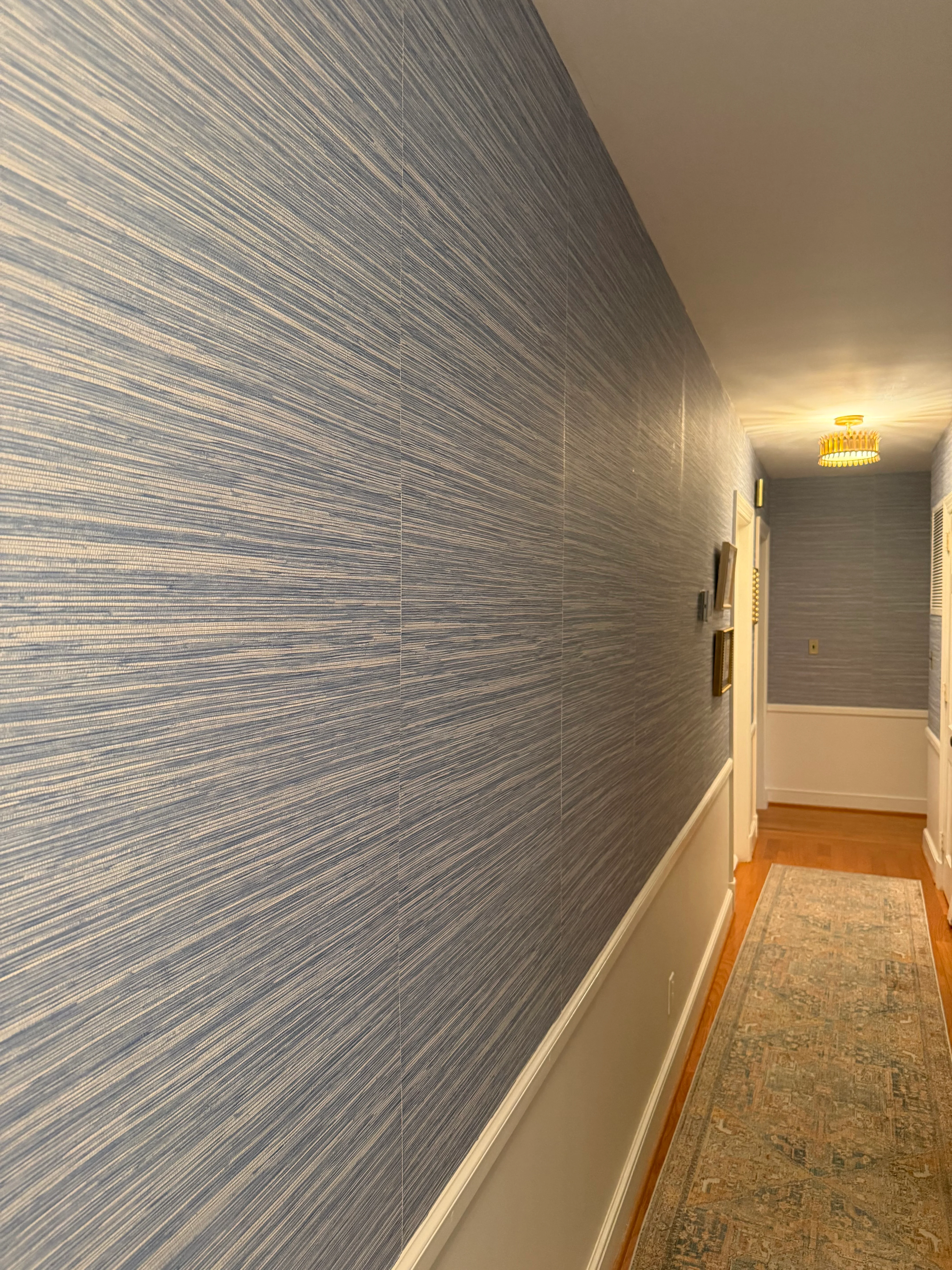 A hallway lined in deep blue vertical grasscloth above wainscoting, with a vintage patterned runner rug on the floor.