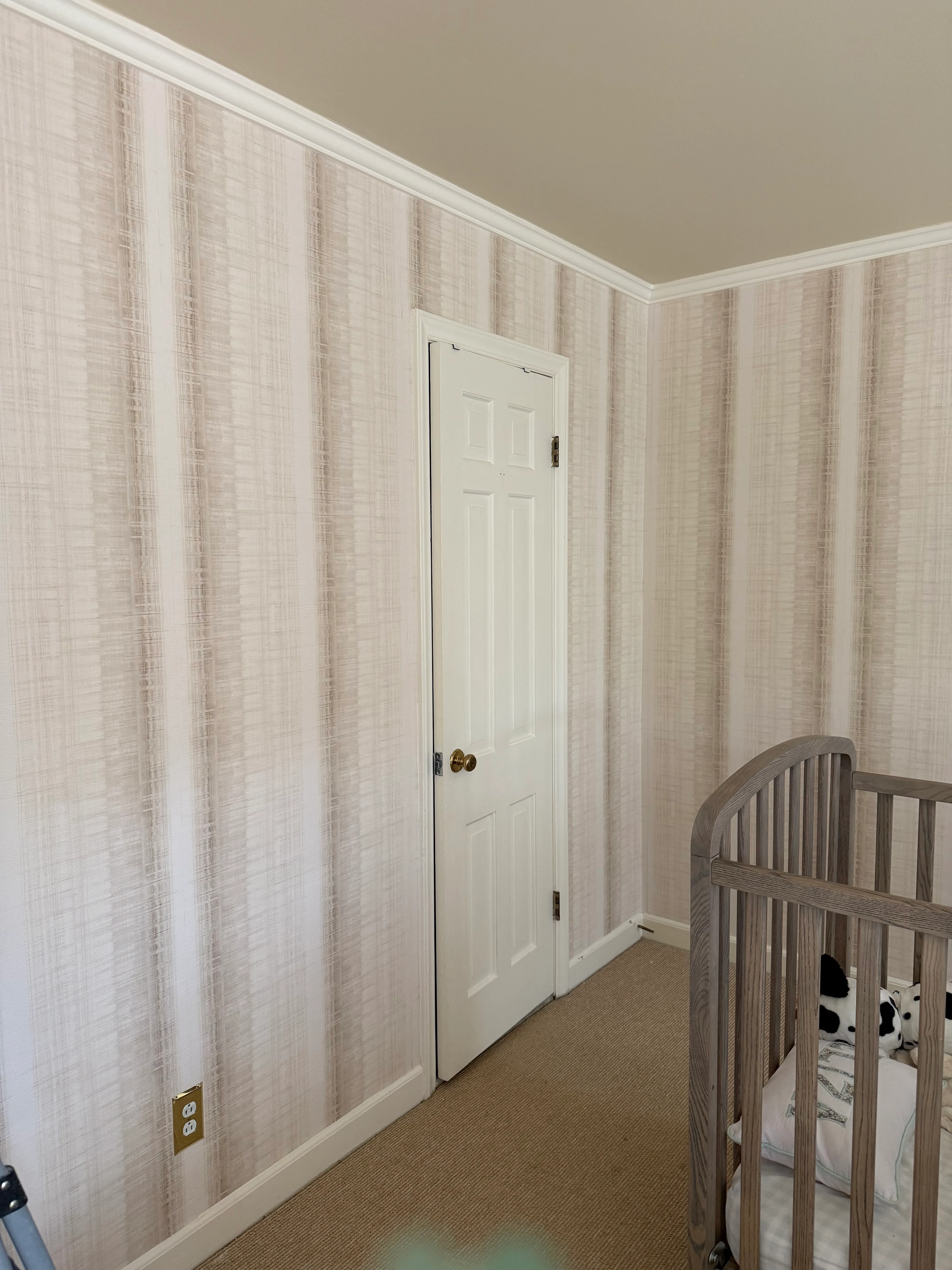 A nursery with soft beige painterly stripe wallpaper on all four walls, with a wooden crib in the corner.